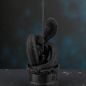 May include: A black cobra incense holder with a detailed, textured design. The snake is coiled, with its head raised, holding an incense stick. Blue smoke rises from the stick, creating a mystical effect. The base features ornate patterns.