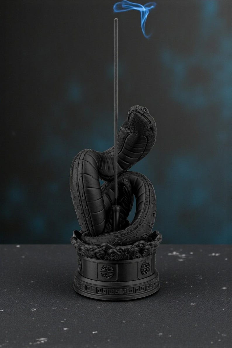 May include: A black cobra incense holder with a detailed, textured design. The snake is coiled, with its head raised, holding an incense stick. Blue smoke rises from the stick, creating a mystical effect. The base features ornate patterns.