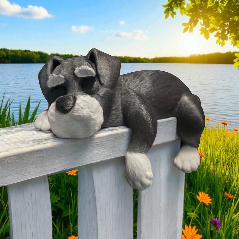 May include: A gray and white dog statue resting on a white wooden bench. The dog has a relaxed pose with its paws hanging over the side. The background features a lake, blue sky, and green foliage.
