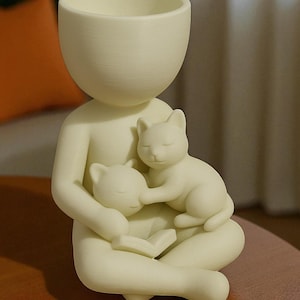 May include: A cream-coloured ceramic planter in the form of a person cradling two sleeping cats. The person is seated with a book, and the planter's head is a large, open bowl. The cats are nestled in the person's arms.