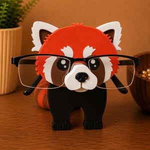May include: A red panda-shaped eyeglasses holder with black-framed glasses. The holder features a red body, white accents, and a black base. The glasses are positioned as if the panda is wearing them, creating a whimsical and functional desk accessory.