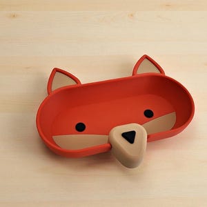 May include: A red fox-shaped tray with a beige snout and ears, and black eyes. The tray is made of a smooth material and sits on a light wood surface. The tray is designed to hold small items.