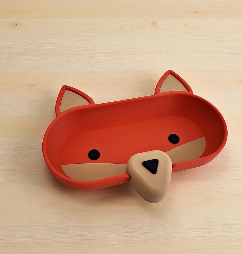 May include: A red fox-shaped tray with a beige snout and ears, and black eyes. The tray is made of a smooth material and sits on a light wood surface. The tray is designed to hold small items.