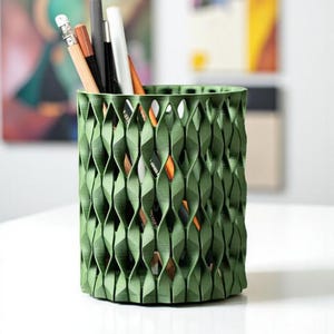 May include: A green, cylindrical desk organizer with a woven, geometric design. The organizer holds various pencils and pens, including a pencil with a white eraser, a black pen, and a white pen. The organizer is on a white surface.