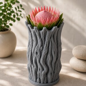 Op de afbeelding: Een grijze, getextureerde vaas met een boomschorsontwerp, met een roze protea-bloem met groene bladeren. De vaas heeft een cilindrische vorm en staat op een lichtgekleurd oppervlak. Een potplant en twee gladde stenen staan op de achtergrond.