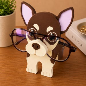 Puede incluir: Un soporte para gafas con forma de chihuahua marr&oacute;n y blanco. El soporte tiene orejas grandes con interiores rosas y sostiene un par de gafas. Las gafas tienen una montura marr&oacute;n oscuro.