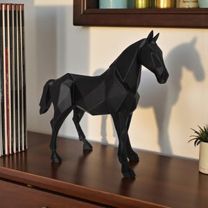 May include: A black geometric horse sculpture with a faceted design stands on a wooden surface. The sculpture is a modern decorative piece, with a shadow cast on the wall behind it. A small potted plant is visible on the right.