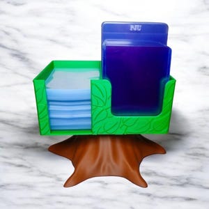 May include: A green and brown card holder with a tree trunk base. The holder has two compartments, one for cards and one for a blue card holder. The green section has a leaf design. The blue card holder has the letters "PJU".