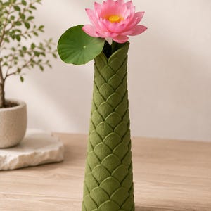 May include: A green, textured vase with a lotus flower and leaf. The vase has a unique scale-like pattern. The pink lotus flower has a yellow center. The green leaf is round. The vase is on a wooden surface.