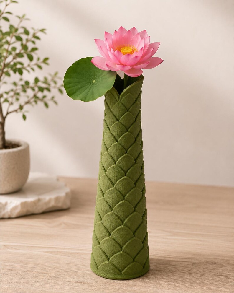 May include: A green, textured vase with a lotus flower and leaf. The vase has a unique scale-like pattern. The pink lotus flower has a yellow center. The green leaf is round. The vase is on a wooden surface.
