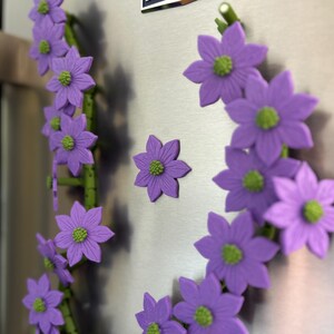 May include: A decorative floral magnet set featuring purple flowers with green centers. The flowers are arranged on green stems, creating a whimsical design. The magnets are attached to a stainless steel surface.