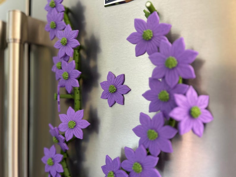 May include: A decorative floral magnet set featuring purple flowers with green centers. The flowers are arranged on green stems, creating a whimsical design. The magnets are attached to a stainless steel surface.
