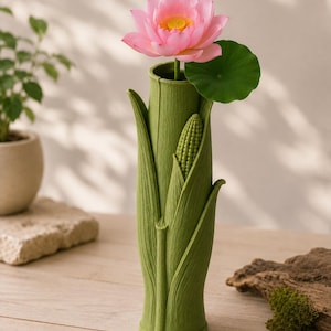 Pu&ograve; includere: Un vaso di ceramica verde a forma di steli di mais, che contiene un fiore di loto rosa con un centro giallo e una foglia verde. Il vaso ha una superficie strutturata e un piccolo dettaglio di pannocchia. L'arrangiamento &egrave; su un tavolo di legno.