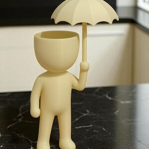 May include: A cream-colored figurine of a person holding a small umbrella. The figurine has a rounded head with a hollowed-out top, and the umbrella is positioned above the head. The figure is standing on a black marble-patterned surface.