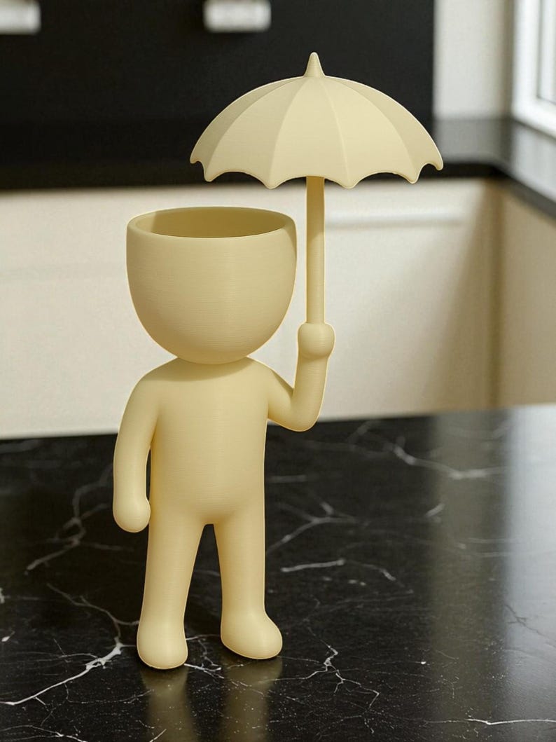 May include: A cream-colored figurine of a person holding a small umbrella. The figurine has a rounded head with a hollowed-out top, and the umbrella is positioned above the head. The figure is standing on a black marble-patterned surface.