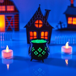May include: A black cauldron filled with green liquid, surrounded by three small white tea lights on a blue surface. The cauldron is in front of three paper Halloween houses with orange lights inside.