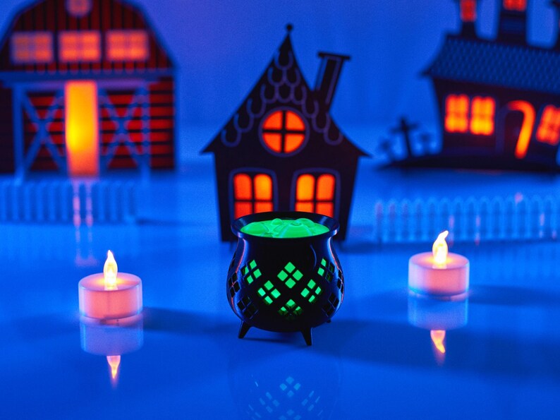 May include: A black cauldron filled with green liquid, surrounded by three small white tea lights on a blue surface. The cauldron is in front of three paper Halloween houses with orange lights inside.