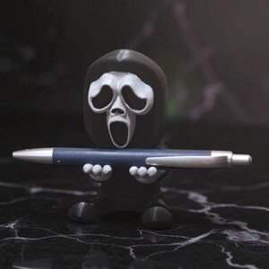 Puede incluir: Una figura negra de Ghostface, el asesino de Scream, sosteniendo un bol&iacute;grafo azul con detalles plateados. La figura tiene una m&aacute;scara blanca con la boca abierta. Est&aacute; sobre una superficie negra.