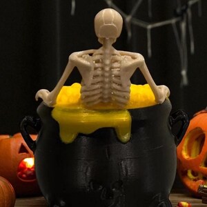 May include: A black cauldron with a yellow liquid and a small skeleton figure. The Halloween-themed decoration also features two carved pumpkins and candy corn.