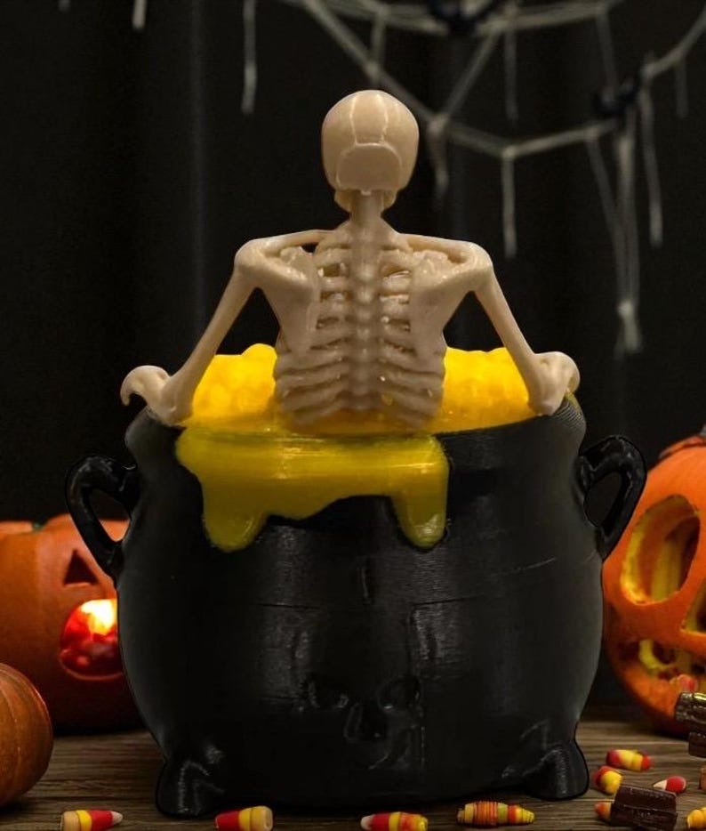 May include: A black cauldron with a yellow liquid and a small skeleton figure. The Halloween-themed decoration also features two carved pumpkins and candy corn.