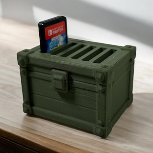 May include: A dark green, 3D-printed chest-shaped Nintendo Switch game cartridge holder. The holder has a slot for a game cartridge, with the "Instant Chef" game visible. The chest has a latch and decorative corner accents.