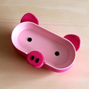 Op de afbeelding: Een roze varkensvormige bak met een rechthoekig lichaam, twee zwarte stipogen en een ronde snuit met twee zwarte stippen. De bak heeft twee roze oren en is gemaakt van plastic.