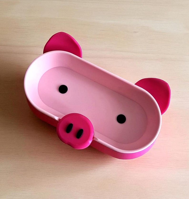 Op de afbeelding: Een roze varkensvormige bak met een rechthoekig lichaam, twee zwarte stipogen en een ronde snuit met twee zwarte stippen. De bak heeft twee roze oren en is gemaakt van plastic.