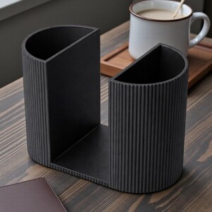 May include: A black desk tidy with a ribbed texture, featuring two semi-circular compartments. A brown leather wallet sits nearby on a wooden surface. A white mug with coffee and a wooden spoon rests on a wooden tray.