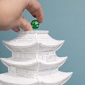May include: A white, multi-tiered pagoda-style structure with intricate architectural details. A green, multi-sided die is held above the pagoda. The die has white numbers and is being dropped into the pagoda.