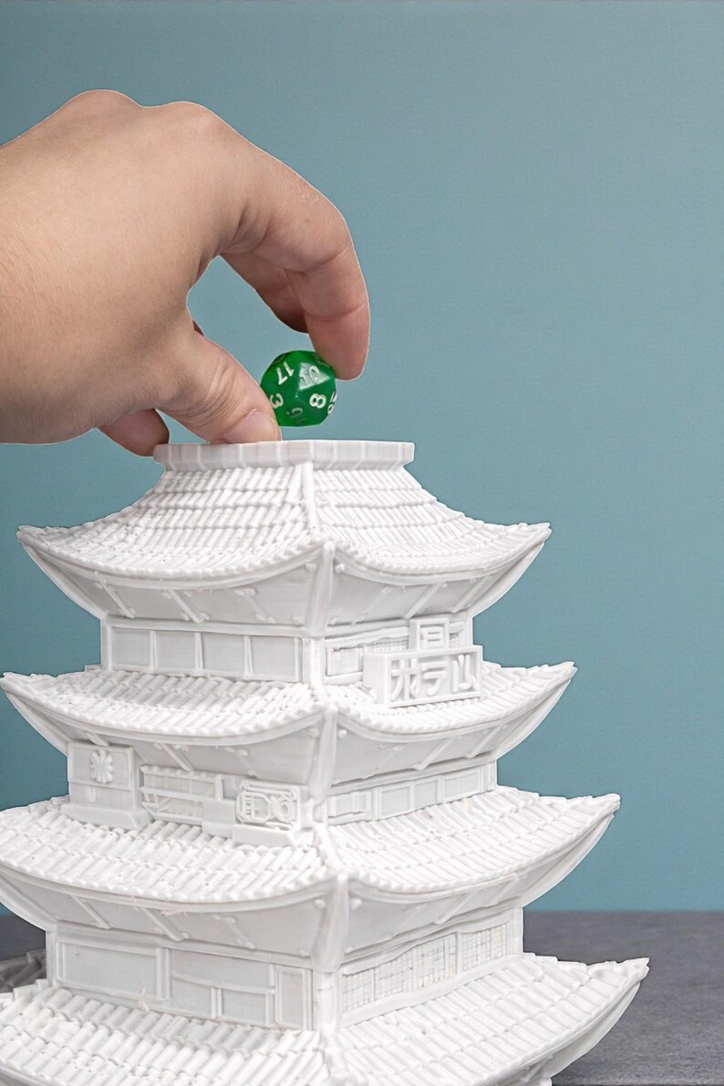 May include: A white, multi-tiered pagoda-style structure with intricate architectural details. A green, multi-sided die is held above the pagoda. The die has white numbers and is being dropped into the pagoda.