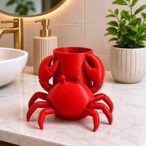 May include: A bright red crab-shaped container, likely a toothbrush holder, sits on a marble countertop. The container has a cylindrical shape with large crab claws on either side. A smaller crab is in front of the container.