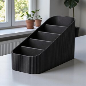 May include: A black desk organizer with a sloped design and four compartments. The organizer has a textured, ribbed exterior and is made of a solid, dark material. It sits on a white surface, with plants visible in the background.