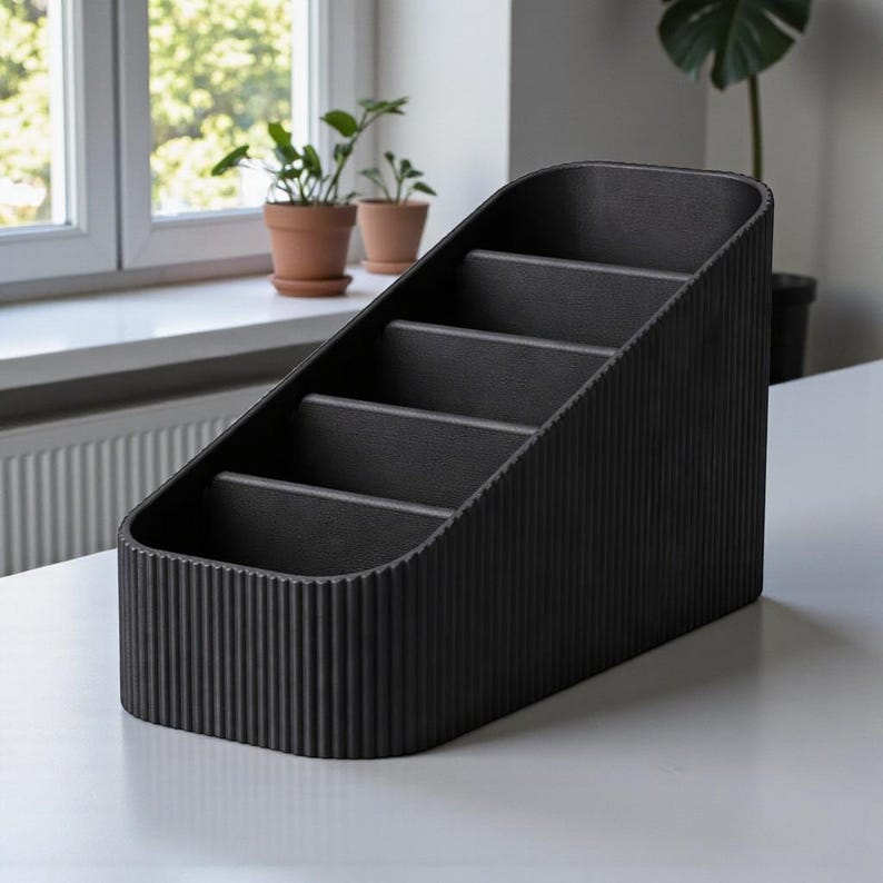 May include: A black desk organizer with a sloped design and four compartments. The organizer has a textured, ribbed exterior and is made of a solid, dark material. It sits on a white surface, with plants visible in the background.