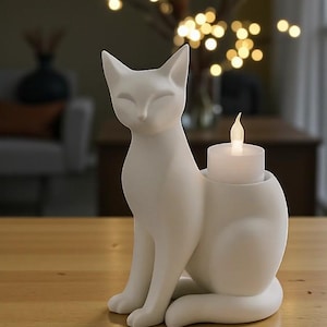 Op de afbeelding: Een witte keramische kandelaar in de vorm van een zittende kat. De kat heeft gesloten ogen en puntige oren. Hij houdt een witte kaars met een flikkerende vlam vast. De kandelaar staat op een houten oppervlak.