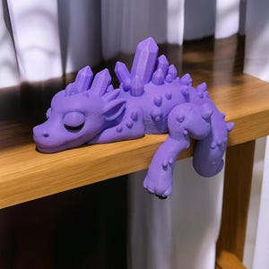 May include: A lavender-colored dragon figurine with crystal-like protrusions along its back and head. The dragon is lying down with its eyes closed and its front leg extended. The figurine is resting on a wooden surface.
