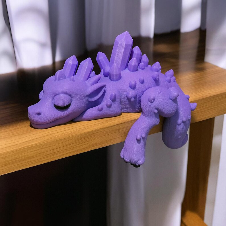 May include: A lavender-colored dragon figurine with crystal-like protrusions along its back and head. The dragon is lying down with its eyes closed and its front leg extended. The figurine is resting on a wooden surface.