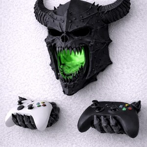 May include: A black, horned skull wall decor with glowing green light emanating from the mouth, above two controller holders. One controller holder is white, the other is black. The controllers have colored buttons.