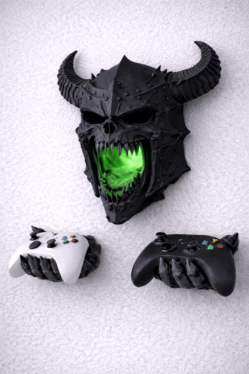 May include: A black, horned skull wall decor with glowing green light emanating from the mouth, above two controller holders. One controller holder is white, the other is black. The controllers have colored buttons.