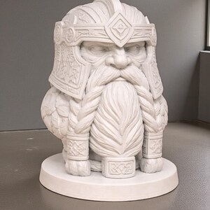 May include: A white sculpture of a dwarf's head and upper body, featuring a long, braided beard and a detailed helmet. The statue is on a circular base and has a stern expression. The dwarf is a decorative item.