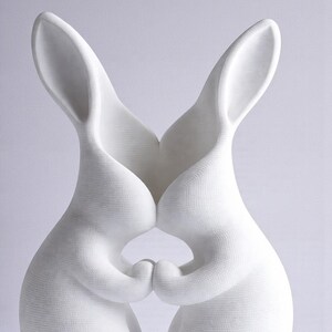 May include: A white ceramic sculpture of two rabbits in a loving embrace. The rabbits are touching noses and holding hands, creating a heart shape. The sculpture has a textured surface and is set against a light background.