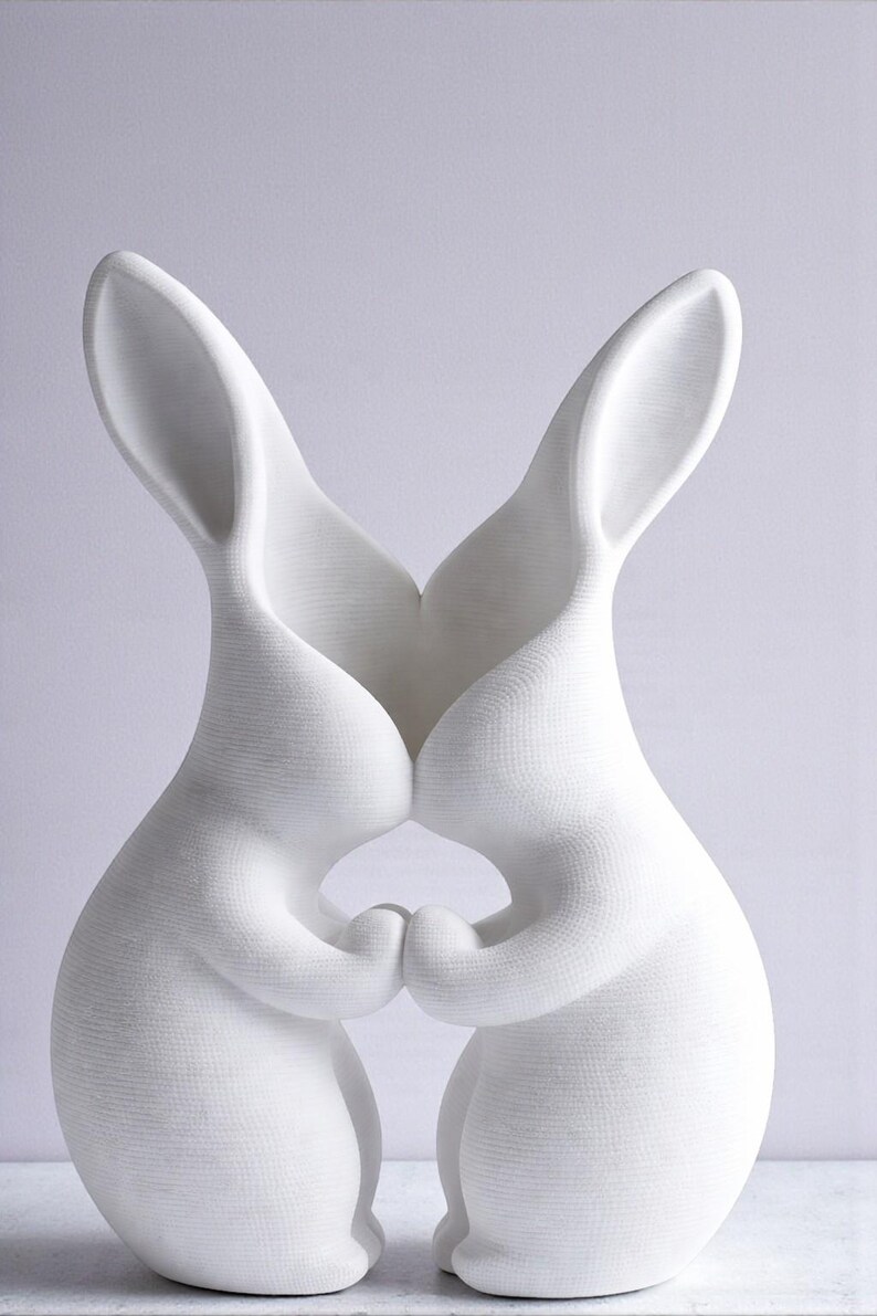 May include: A white ceramic sculpture of two rabbits in a loving embrace. The rabbits are touching noses and holding hands, creating a heart shape. The sculpture has a textured surface and is set against a light background.