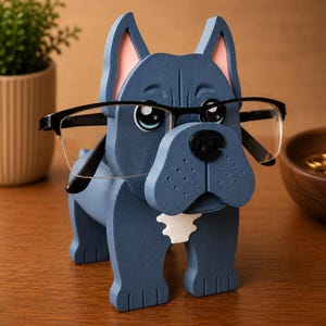 May include: A blue wooden dog-shaped glasses holder with black-framed glasses. The dog has pink inner ears and a white patch on its chest. The holder is on a wooden surface, with a small plant and a wooden bowl in the background.