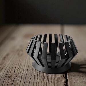 Puede incluir: Un portavelas geom&eacute;trico negro con base circular y listones verticales. El portavelas es de metal y se asienta sobre una superficie de madera. El dise&ntilde;o es moderno y minimalista.