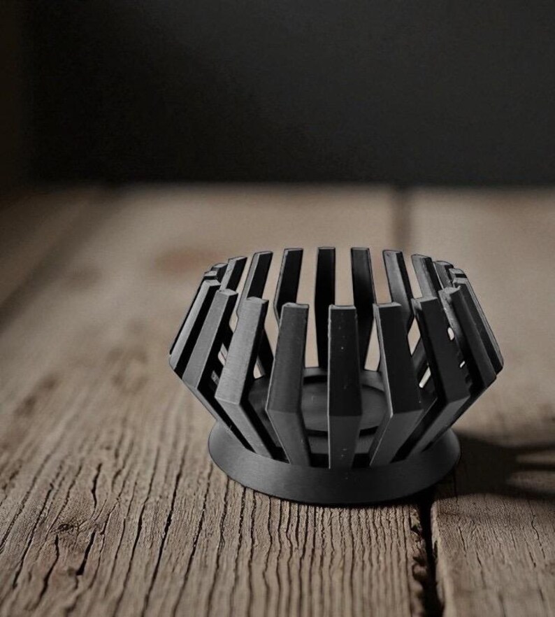 Puede incluir: Un portavelas geom&eacute;trico negro con base circular y listones verticales. El portavelas es de metal y se asienta sobre una superficie de madera. El dise&ntilde;o es moderno y minimalista.