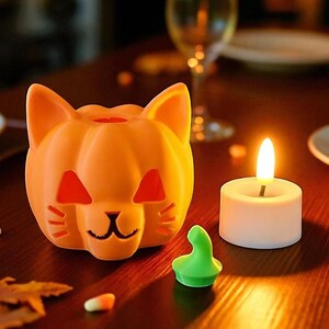 May include: An orange cat-shaped candle holder with cut-out eyes and a mouth, next to a lit white candle. A small green object and candy corn are on a wooden surface. The scene is lit with warm, inviting light.