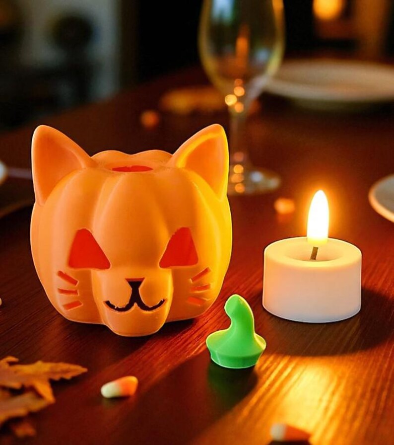 May include: An orange cat-shaped candle holder with cut-out eyes and a mouth, next to a lit white candle. A small green object and candy corn are on a wooden surface. The scene is lit with warm, inviting light.