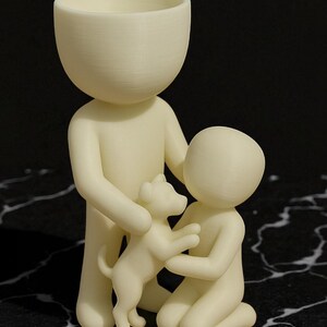 May include: A cream-colored figurine depicting a family with a dog. The tallest figure holds the dog, while a smaller figure kneels, embracing the dog. The figures have rounded heads and simple bodies, creating a minimalist aesthetic.