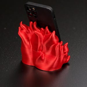 Puede incluir: Un soporte para tel&eacute;fono rojo brillante con forma de llamas, sosteniendo un tel&eacute;fono inteligente negro. El soporte es de un material liso y est&aacute; sobre una superficie oscura. El dise&ntilde;o es moderno y llamativo, para un escritorio o mesita de noche.