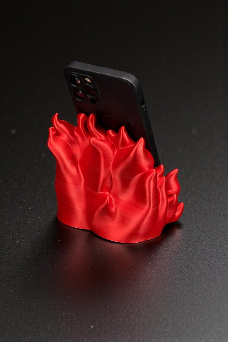 Puede incluir: Un soporte para tel&eacute;fono rojo brillante con forma de llamas, sosteniendo un tel&eacute;fono inteligente negro. El soporte es de un material liso y est&aacute; sobre una superficie oscura. El dise&ntilde;o es moderno y llamativo, para un escritorio o mesita de noche.