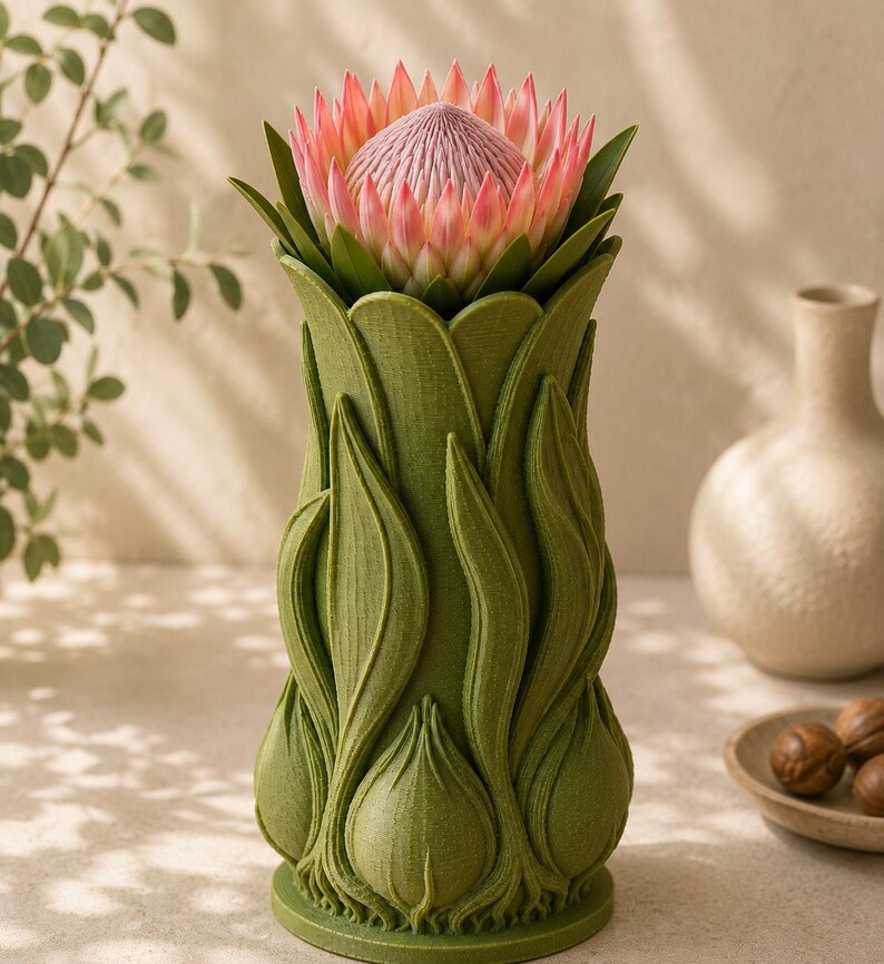 May include: A green vase with a detailed leaf design, holding a large pink and green protea flower. The vase has a textured surface and a unique, organic shape, creating a striking floral arrangement.